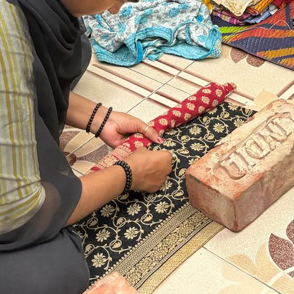 A woman sewing on the floor of the workshop beside folded sari fabric.